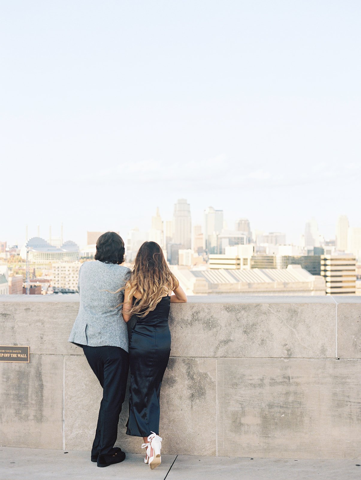 A couple dressed in a suit and a black dress admiring the Kansas City skyline at the Liberty Memorial Tower during their engagement session in Downtown Kansas City, Missouri