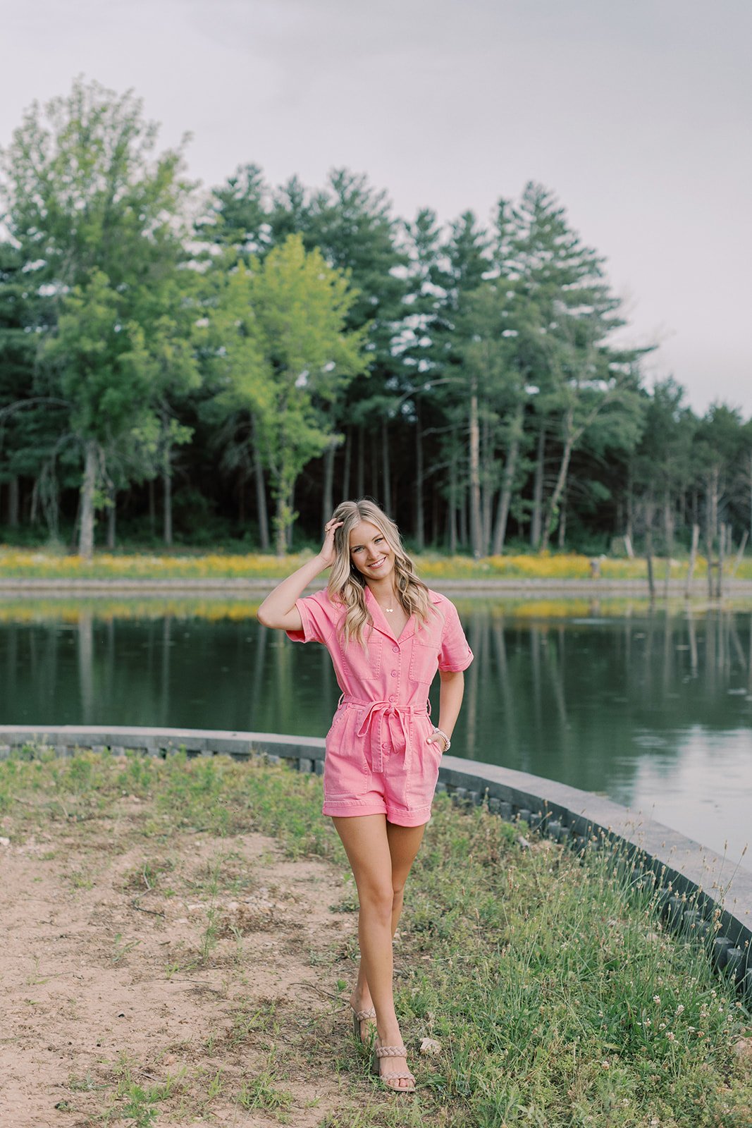 High school senior female standing near water in a hot pink jumpsuit posing for senior pictures
