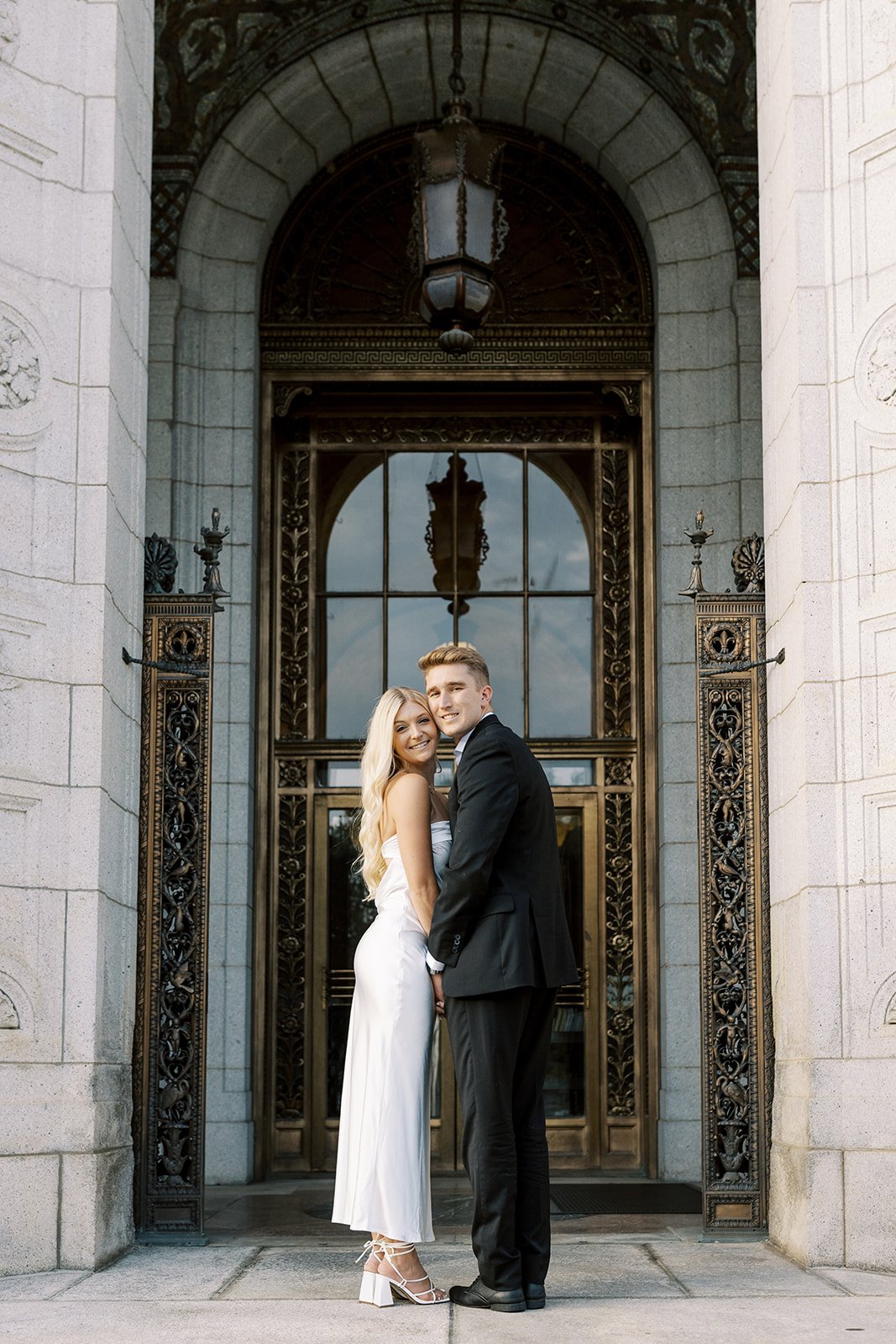A couple dressed in a long white dress and a black suit are posing during their engagement session.